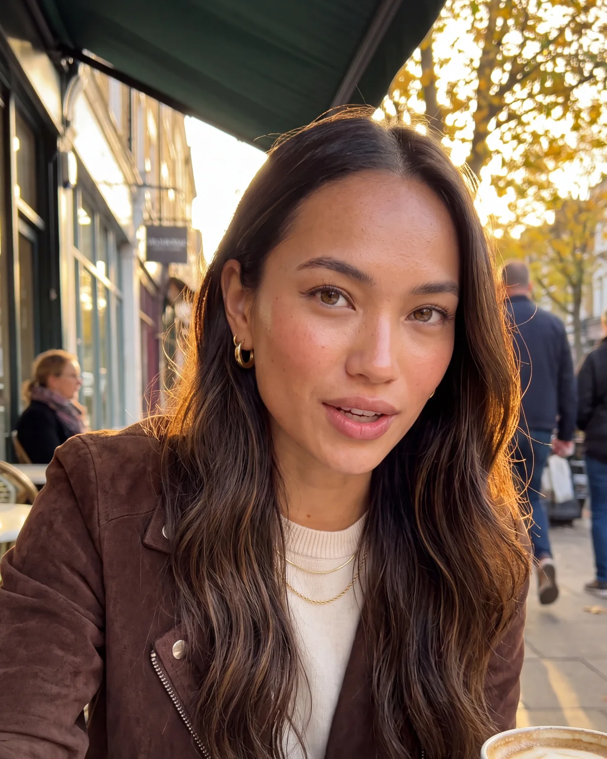 Mia Aquino at a London café table in autumn sun, wearing small gold hoops and a brown suede jacket
