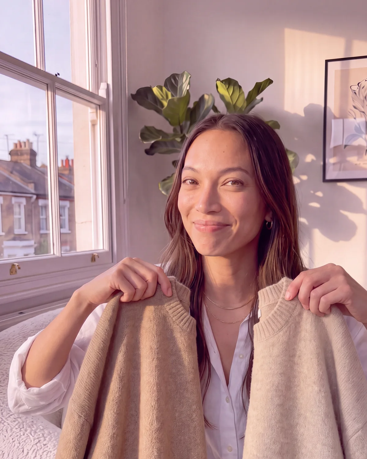 Mia Aquino holding up two camel crew-neck jumpers side by side in a sunny London flat