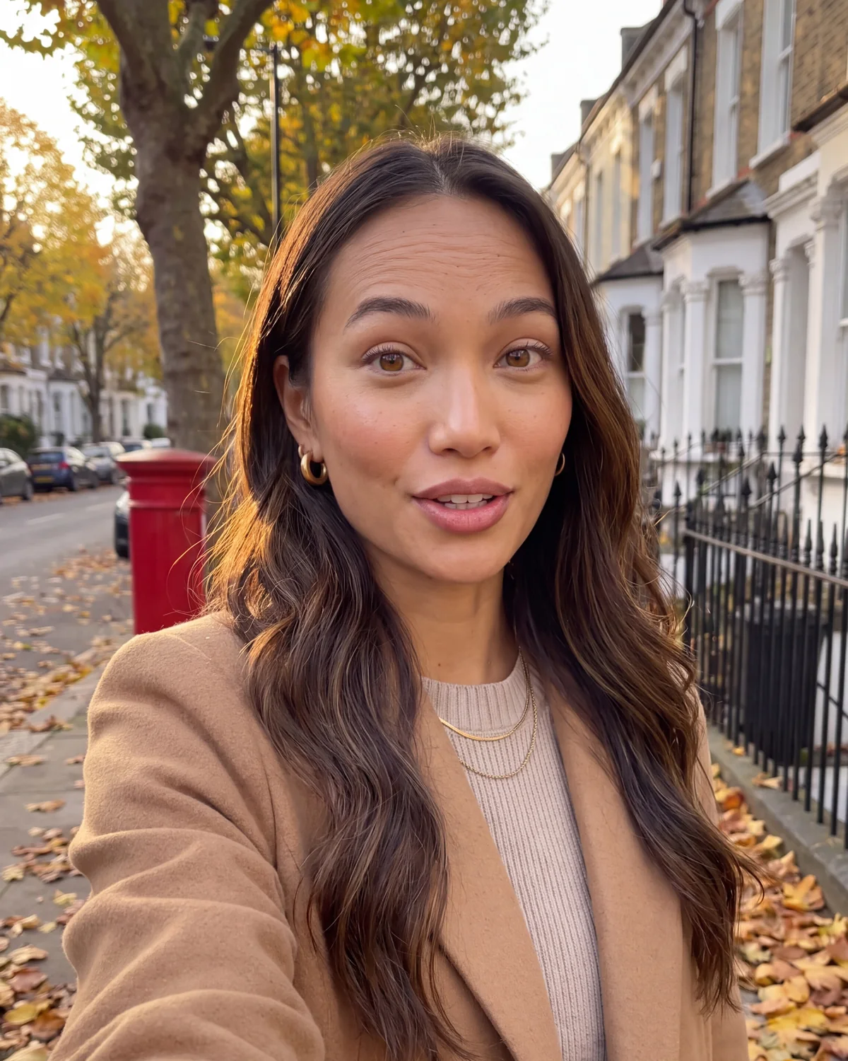 Mia Aquino in a camel coat on a leafy London street in autumn, with a red postbox behind her